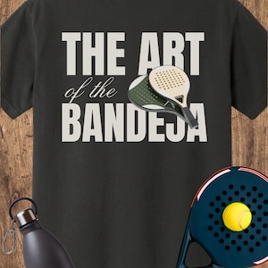 May include: A black t-shirt featuring the white text THE ART of the BANDEJA and an illustration of two padel rackets. A dark blue padel racket with a yellow ball and a black water bottle are positioned in the foreground of the t-shirt design.
