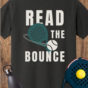 May include: A black t-shirt featuring the white text READ THE BOUNCE with a graphic of a teal padel racket and a white ball. A black water bottle and a dark blue padel racket with a yellow ball are positioned near the shirt.