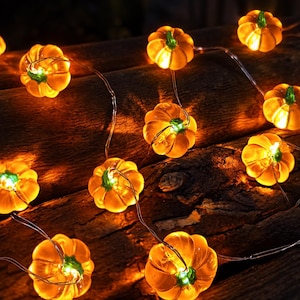 Puede incluir: Una cadena de luces LED con pequeñas cubiertas translúcidas en forma de calabaza de color naranja con tallos verdes. Cada calabaza brilla desde el interior, conectada por un cable plateado fino y flexible que permite una disposición versátil.