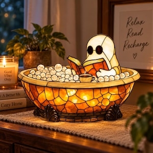 May include: A stained glass lamp shaped like a bathtub filled with white glass bubbles, featuring a ghost figure wearing round glasses and reading an open book. A small cup sits among the bubbles. The base has a warm orange glow and dark metal feet.