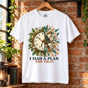 May include: A white short-sleeve t-shirt featuring a graphic of a partially shattered clock surrounded by green leaves and small white flowers. The text "I HAD A PLAN FOR THAT." is printed in black and orange below the clock illustration.