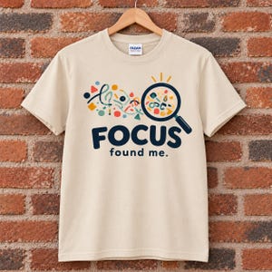 May include: A beige short-sleeved t-shirt featuring a graphic of a magnifying glass over colourful abstract shapes and the text "FOCUS found me." The design includes scattered geometric elements in blue, red, yellow, and green colours on the front.