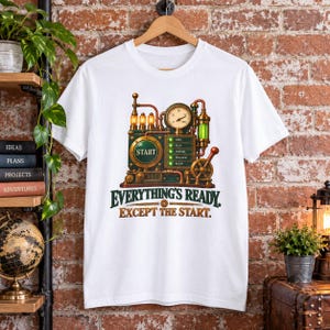 May include: A white short-sleeve t-shirt featuring a graphic of a mechanical device with a pressure gauge, glowing lights, and a large button labeled START. Below the device, the text reads: EVERYTHING'S READY. EXCEPT THE START.