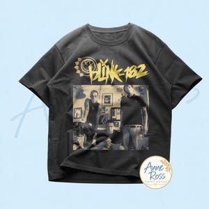 May include: A charcoal gray short-sleeve t-shirt featuring a gold graphic of the Blink-182 logo and band name above a black and white photograph of the three band members standing in a room with framed pictures on the wall.