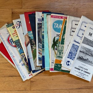 May include: A collection of vintage travel brochures and maps arranged in a fan shape. The documents feature various colors, illustrations, and text including 'Information Guide and Map of Lakeland Florida' and 'Official Map Tampa'.