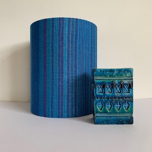 May include: A cylindrical lampshade featuring a vertical striped pattern in shades of blue and purple, paired with a rectangular ceramic object in teal and dark blue with a repeating geometric diamond relief pattern across its front surface.