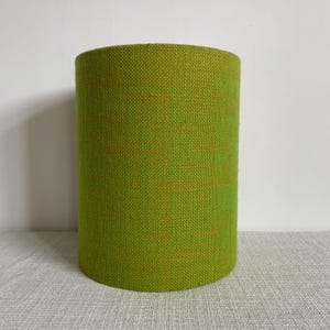 May include: A cylindrical lampshade featuring a vibrant lime green fabric with a visible, coarse-woven texture. The material displays a consistent weave pattern throughout the entire surface, creating a uniform appearance for this drum-shaped lighting accessory.