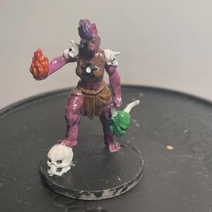 Peut inclure: Une figurine peinte à la peau violette, portant un pagne marron et des épaulettes blanches. Le personnage tient une flamme orange vif dans une main et un masque vert dans l'autre, tout en posant un pied sur un crâne blanc.