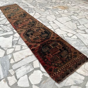 Puede incluir: Una alfombra de pasillo larga y estrecha con un patrón repetitivo de medallones octogonales geométricos en azul marino oscuro y negro sobre un fondo cálido de color rojo óxido y marrón desgastado, con flecos cortos oscuros en los extremos.