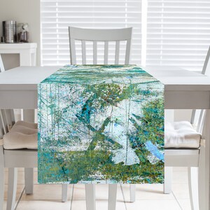 May include: A rectangular table runner with an abstract design in shades of teal, green, and white. The pattern appears to be a textured, painted design, with brushstrokes and splatters. The runner is displayed on a white table.