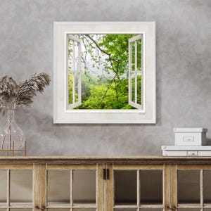Puede incluir: Una obra de arte enmarcada que representa una ventana abierta con vistas a un exuberante bosque verde. El marco blanco de la ventana contrasta con el vibrante follaje verde y el fondo brumoso. La obra está montada en una pared gris.