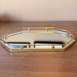 以下が含まれることがあります： ゴールドトーンの金属フレームが付いた八角形のミラートレイ。ベースの縁には複雑なエンボス加工のフローラルパターンが施されており、盛り上がった手すりは八角形の各コーナーにある小さな球状のアクセントによって支えられています。