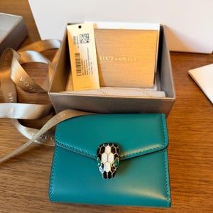 Puede incluir: Una cartera compacta de cuero color verde azulado con un cierre de cabeza de serpiente con detalles en esmalte negro, blanco y verde. La cartera se presenta en una caja dorada de marca con una cinta beige y una etiqueta con el nombre Bvlgari.