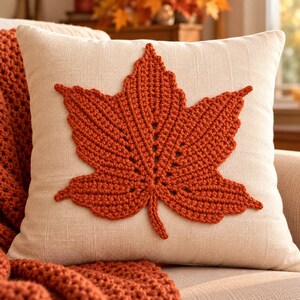 May include: A square throw pillow with a cream-colored fabric cover featuring a large, centered, burnt-orange crocheted maple leaf appliqué. The leaf has detailed, textured stitching that mimics the veins of a natural leaf, centered on the pillow.