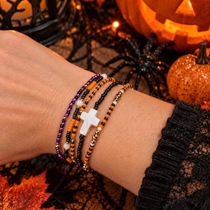 Peut inclure: Un ensemble de quatre bracelets de perles aux couleurs noir, orange, violet et or métallique. Les bracelets sont composés de petites perles rondes et d'une croix blanche centrale, avec quelques brins intégrant de petites perles blanches.