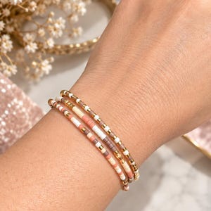 Peut inclure: Un ensemble de trois bracelets de perles délicats composés d'un motif répétitif de petites perles cylindriques dans des tons blanc, pêche et brun sourd, entrecoupées de minuscules perles intercalaires dorées sur un cordon élastique.