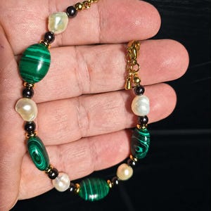 Peut inclure: Un bracelet composé d'une alternance de perles ovales en malachite verte, de perles baroques blanches et de petites perles métalliques sombres, reliées par des entretoises dorées et un fermoir mousqueton doré.