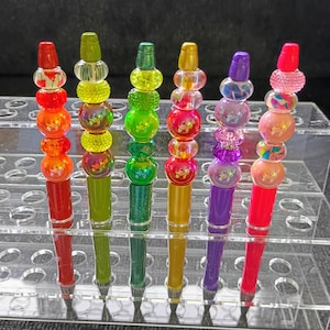 Puede incluir: Seis bolígrafos dispuestos en un soporte de exhibición de acrílico transparente. Cada bolígrafo presenta un color de cuerpo único y está rematado con una pila de cuentas decorativas de diversas formas, texturas y colores, incluyendo rojo, verde lima, esmeralda, oro, morado y rosa.