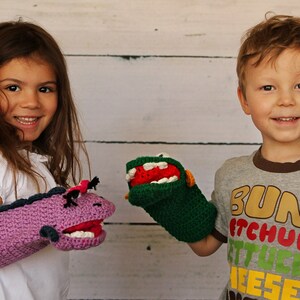 May include: Two children are holding hand puppets. The girl on the left is holding a purple and pink puppet with a white mouth and black eyes. The boy on the right is holding a green puppet with a red mouth and white teeth. The boy is wearing a grey t-shirt with the words "Bun, Ketchup, Lettuce, Cheese" printed on it.