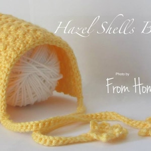 Puede incluir: Un gorro de bebé de crochet amarillo con un patrón de punto de concha. El gorro tiene una larga corbata y dos pequeñas flores de crochet en los extremos de la corbata. El texto "Hazel Shells Bonnet" está escrito en cursiva blanca sobre el gorro. El texto "Photo by From Home" está escrito en blanco debajo del gorro.