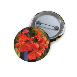 May include: A circular pin-back button featuring a vibrant cluster of bright orange geranium flowers in full bloom. The back of the button is made of silver-toned metal and includes a horizontal pin mechanism for fastening to fabric or clothing.