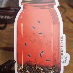 May include: A red glass jar with a white lid filled with a variety of colorful flies. The jar has the text "STICKY FLY CO." written on the side. The jar is on a white background.