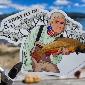 Könnte beinhalten: Ein Aufkleber mit einer Illustration von Thomas Jefferson, der einen Fisch hält. Der Aufkleber trägt den Text "STICKY FLY CO." und "We the People". Der Fisch ist gold und rot. Jefferson trägt ein grünes Hemd und einen rot-weiß-blauen Schal.