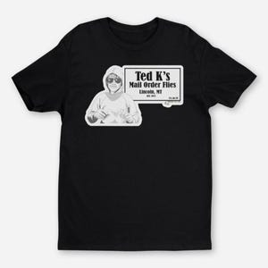 May include: Black t-shirt with a white graphic of a person tying a fishing fly. The graphic includes the text "Ted K's Mail Order Flies, Lincoln, MT, Est. 1973."