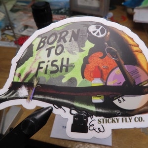 May include: A green, brown, and orange camouflage sticker of a military helmet with the text "Born to Fish" and a peace sign. The sticker also features a fly fishing lure, a bottle of fly tying thread, and a compact disc with the text "Sticky Fly Co."