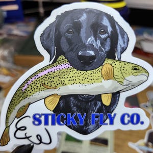 以下が含まれることがあります： 黒いラブラドールレトリバーが口にくわえたマスを描いたステッカー。マスは黄色と緑色でピンクの縞模様があります。「STICKY FLY CO.」の文字が青色で書かれています。
