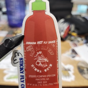 Könnte beinhalten: Ein roter und weißer Aufkleber für eine scharfe Sauce mit grünem Deckel. Das Etikett lautet "Sriracha Hot Fly Sauce" und "Eat,  Chai, Tay". Der Aufkleber ist für eine Firma namens "Sticky Fly Co."