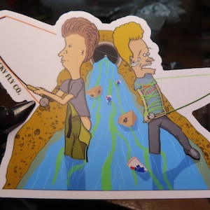 May include: A sticker featuring two cartoon characters fishing in a sewer. The characters are from the animated series "Beavis and Butt-Head". The sticker has the text "Sticky Fly Co." on it.