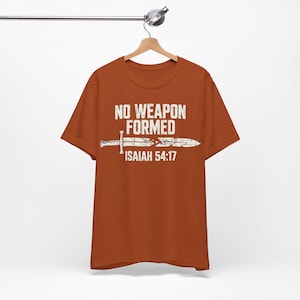 May include: A burnt orange short-sleeve t-shirt featuring white distressed text that reads NO WEAPON FORMED and ISAIAH 54:17, with a graphic of a broken sword positioned horizontally between the two lines of text on the front of the shirt.