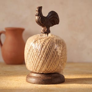 Puede incluir: Un portacuerdas de hierro fundido marrón con un remate en forma de gallo en la parte superior. La base circular sostiene un ovillo de cuerda color canela claro envuelto en plástico transparente. El metal tiene un acabado oscuro y texturizado.