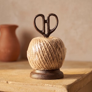 May include: A ball of light brown twine wrapped in clear plastic, resting on a dark brown circular metal base. A decorative dark brown metal scissor-shaped handle extends vertically from the top centre of the twine ball.