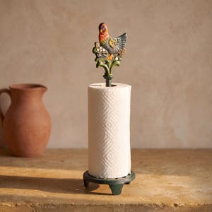 Puede incluir: Un portarrollos de cocina de hierro fundido con una varilla vertical rematada por una figura de metal pintado de una gallina posada sobre una rama verde. La base es un soporte circular verde de tres patas con un rollo de papel blanco.