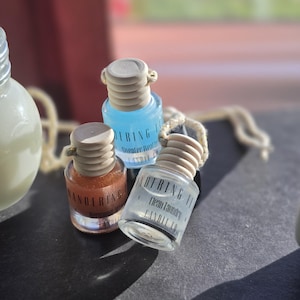 May include: Three small glass car air freshener bottles with wooden caps and hanging cords. The bottles contain blue, orange, and clear liquid. Each bottle features a label with the text Wandering Wick Candle Co and specific scent names.