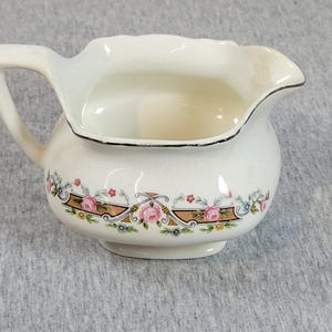 May include: A cream-colored ceramic creamer pitcher featuring a scalloped rim with a silver-toned edge. The body is decorated with a horizontal band of pink roses, green foliage, and small blue and yellow flowers set against a tan geometric border.