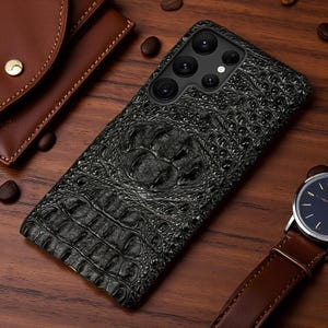 Op de afbeelding: Een zwarte smartphonehoes met een getextureerd, in reliëf aangebracht krokodillenleerpatroon. De hoes heeft een uitsparing voor de camera aan de achterzijde, bestaande uit vier ronde lenzen en een kleine flitser in een verticale opstelling.