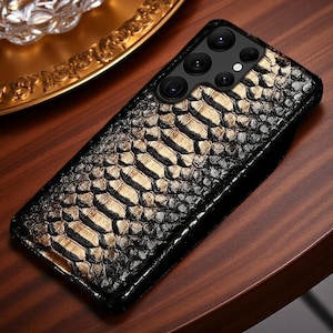 May include: A rectangular smartphone case featuring a textured black and metallic gold snakeskin pattern. The case has a cutout for a multi-lens camera array, showing four circular lenses and a small flash, with a glossy finish throughout.
