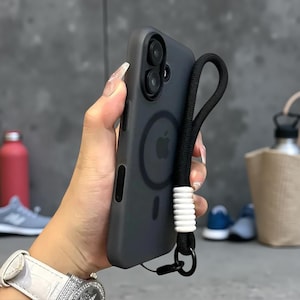 May include: A matte charcoal gray smartphone case with a circular magnetic charging ring and a central Apple logo. The case features a black braided wrist strap attached to the side, accented with a series of white cylindrical beads.