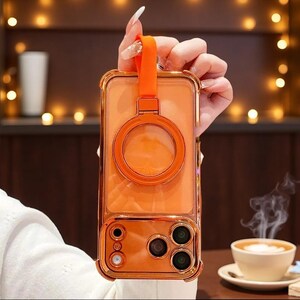 May include: A transparent orange phone case with a metallic orange frame, featuring a circular ring stand on the back and an integrated orange silicone finger strap at the top. The case includes precise cutouts for the camera lenses and flash.