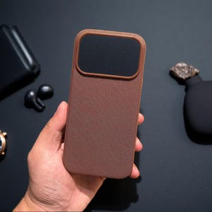 Puede incluir: Una funda rectangular para smartphone con un acabado de tela marrón texturizada y una zona de recorte rectangular negra y lisa para la cámara en la parte superior. La funda presenta esquinas redondeadas y un patrón tejido uniforme.