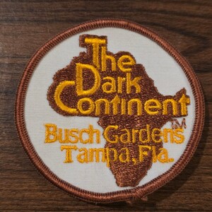 May include: A circular embroidered patch with a white background and a brown border. It features a brown silhouette of the African continent with yellow embroidered text that reads The Dark Continent Busch Gardens Tampa, Fla. TM.