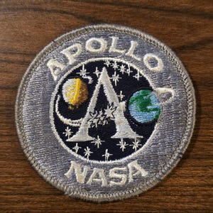 Può includere: Una toppa ricamata circolare con sfondo blu scuro, una grande lettera A bianca, una luna gialla e una Terra blu e verde. Il testo APOLLO è arcuato in alto e NASA è in basso in filo bianco con un bordo argentato.