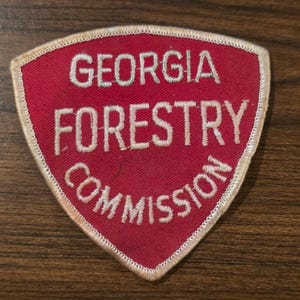May include: A shield-shaped embroidered patch with a red fabric background and a cream-colored border. The text GEORGIA FORESTRY COMMISSION is stitched in cream-colored thread across the center of the patch in three distinct horizontal lines.