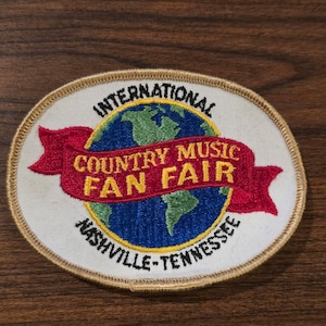 May include: An oval embroidered patch featuring a blue and green globe graphic. A red banner across the center displays the yellow text "COUNTRY MUSIC FAN FAIR". The text "INTERNATIONAL" is arched above, and "NASHVILLE-TENNESSEE" is below.