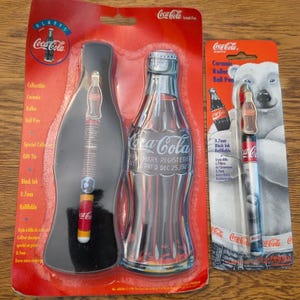 May include: Two Coca-Cola branded writing instruments in original packaging. One is a ceramic rollerball pen inside a bottle-shaped tin. The other is a ceramic rollerball pen on a card featuring a polar bear, with 0.7mm black ink refillable text.