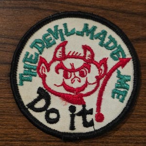 May include: A circular embroidered patch with a cream-coloured background and a black stitched border. It features a red cartoon devil face in the centre, with the green text THE DEVIL MADE ME and the black text Do it stitched around the graphic.