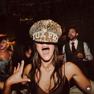 May include: A gold-coloured hat with the text "TUX FIZ" in silver letters, decorated with pearls and rhinestones. The hat is worn by a person with their mouth open, surrounded by other people at a party.
